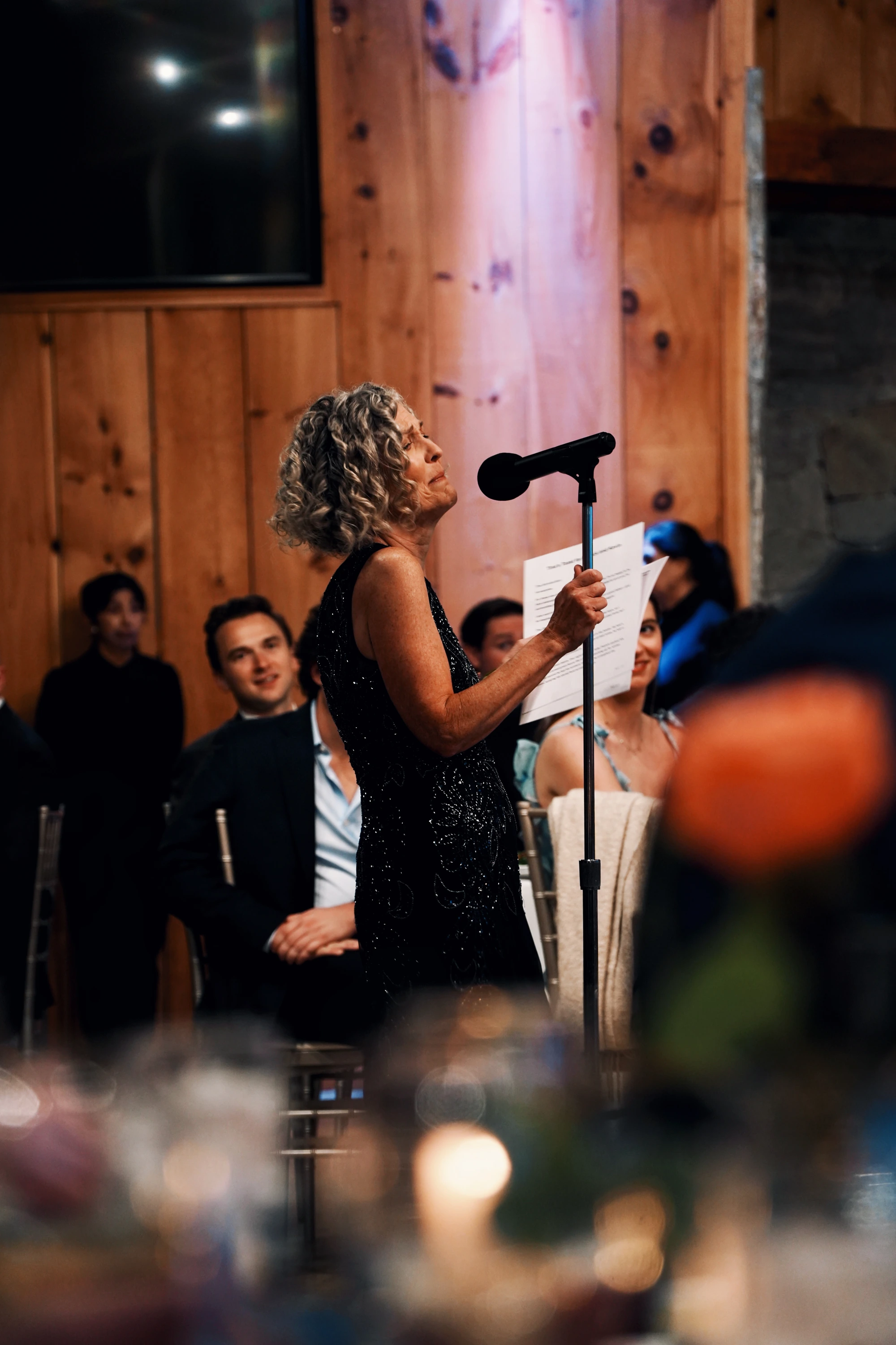 A woman speaks into a microphone among seated wedding guests.