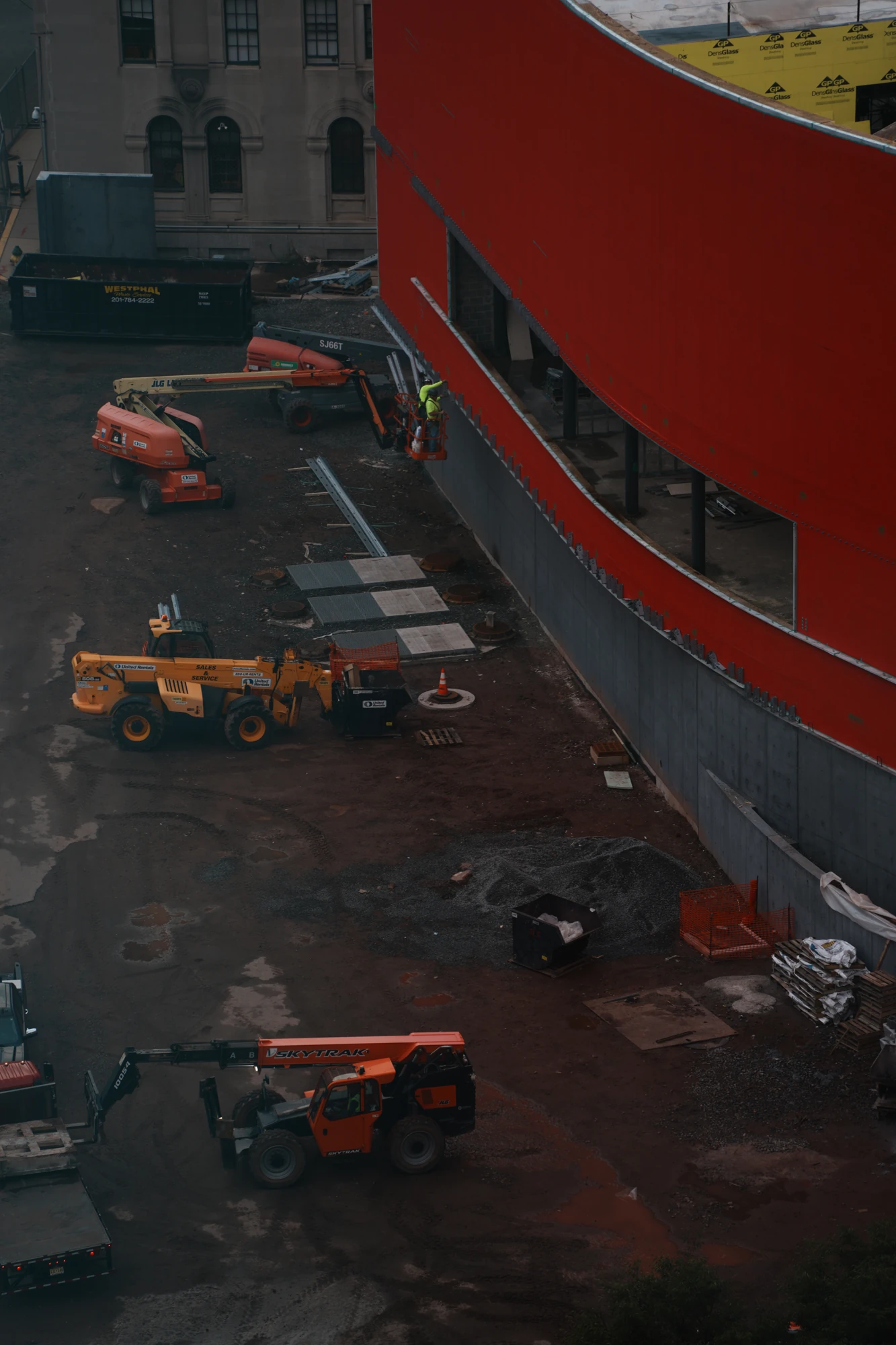Construction vehicles beside the red curve of a building, seen from above.