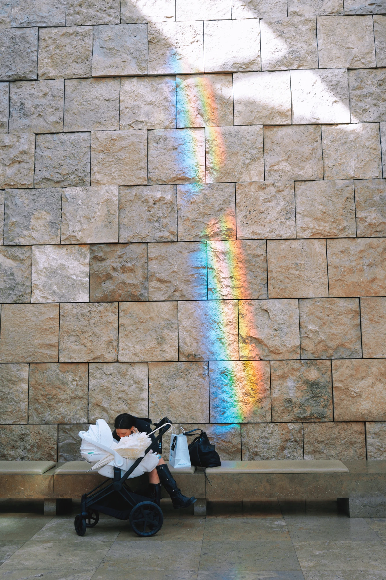A seated visitor and stroller beneath a rainbow of light on a stone wall.