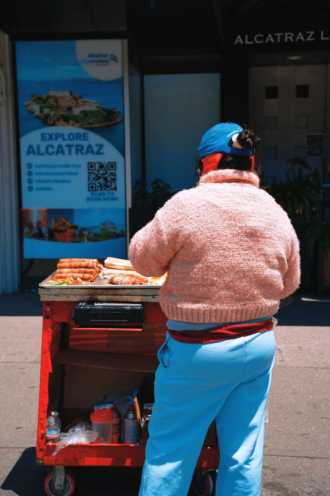 A vendor in a pink jacket tends a cart outside the Alcatraz ferry terminal.