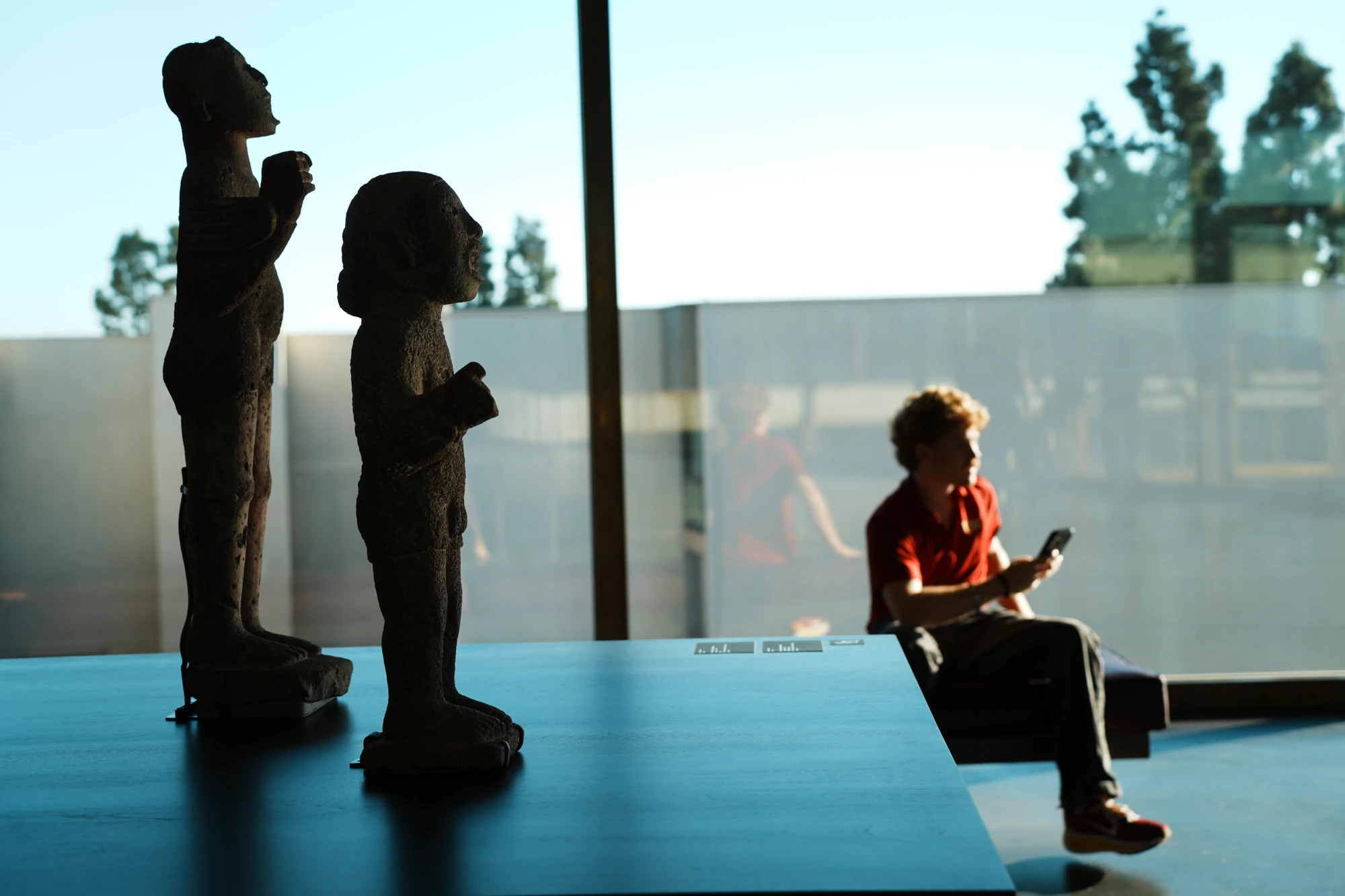 Sculptures stand in silhouette beside a seated visitor at a bright window.