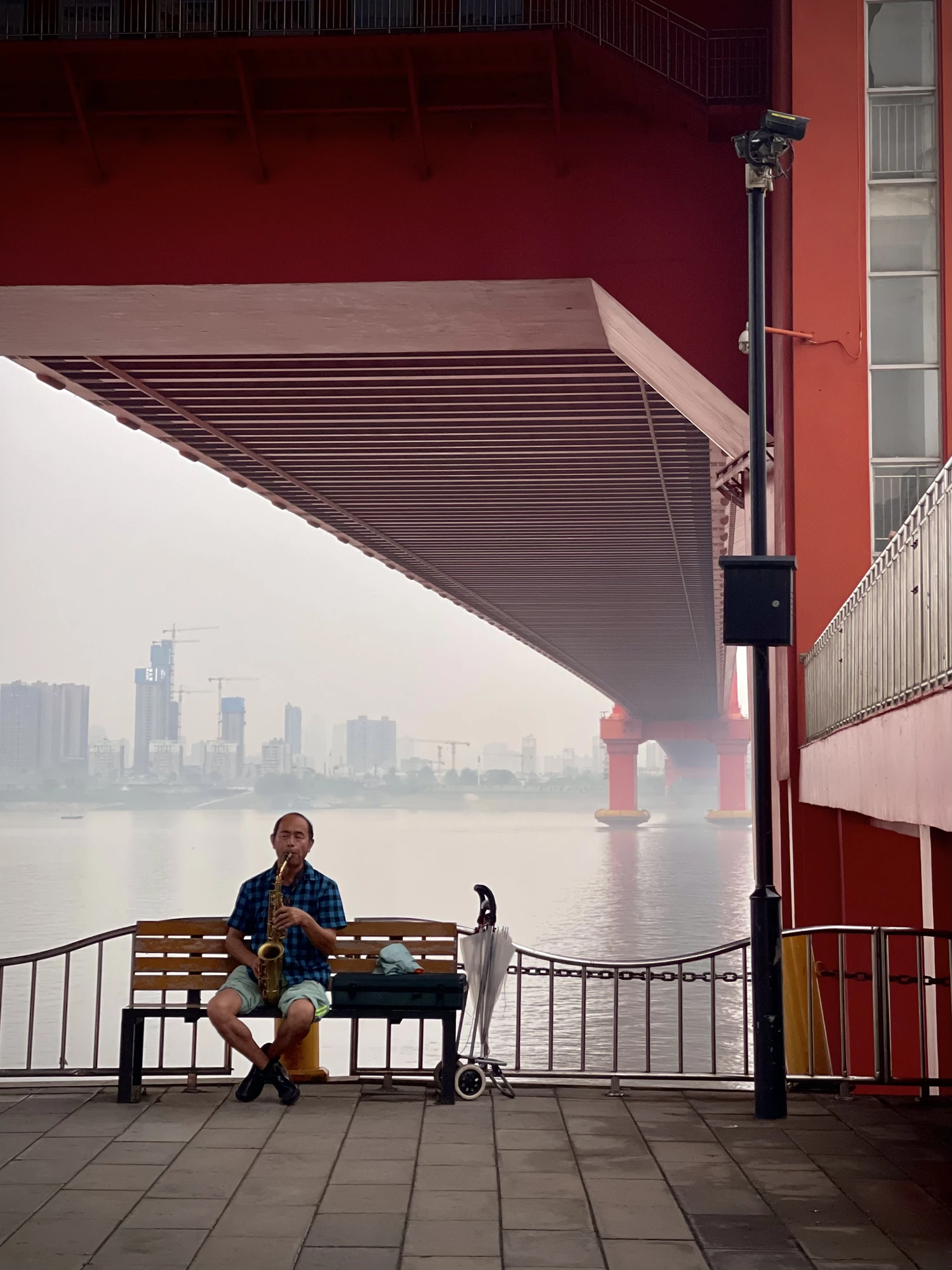A seated saxophonist plays beneath the red underside of a bridge.