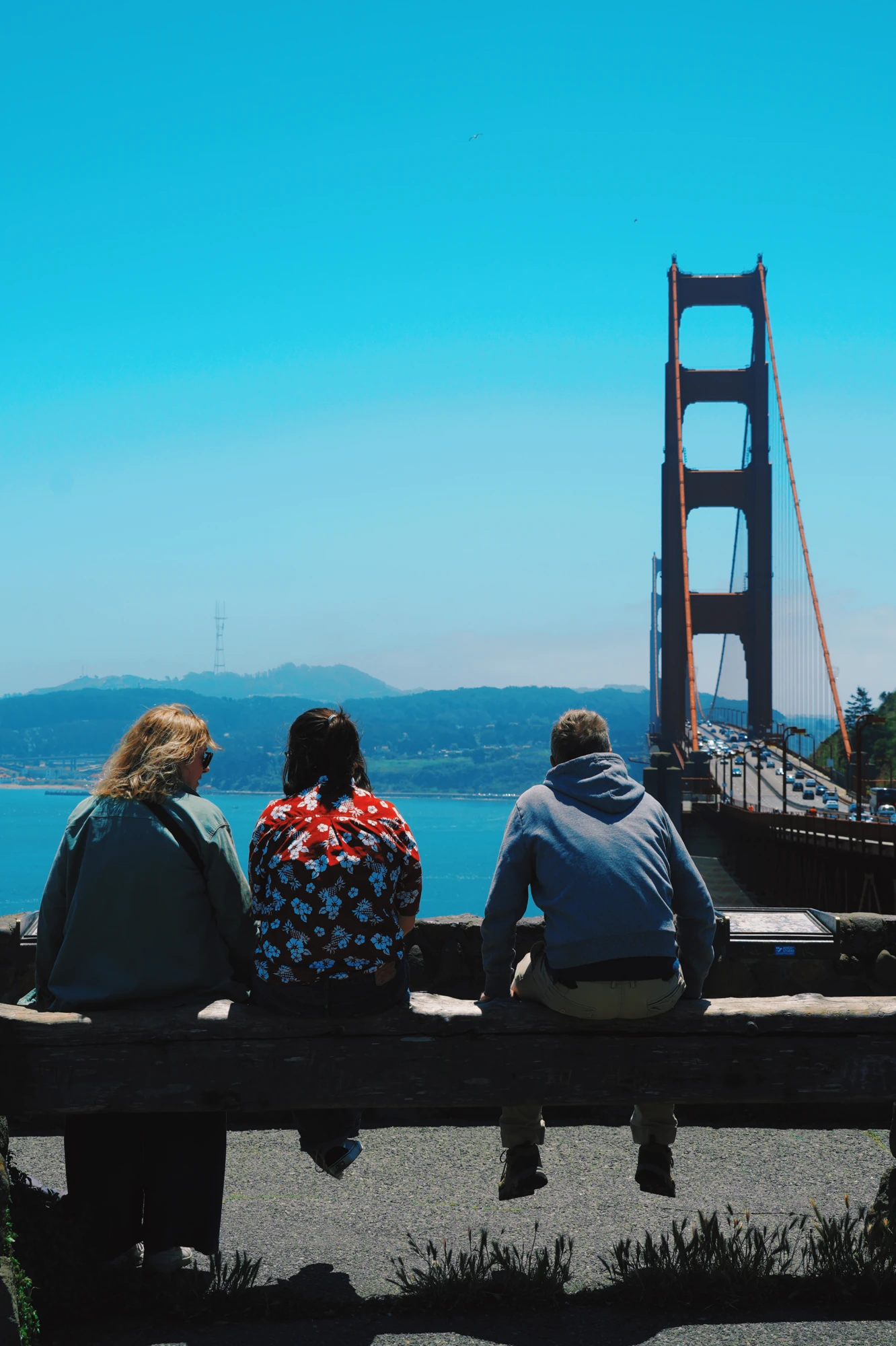 Three people sit on a bench facing the Golden Gate Bridge.