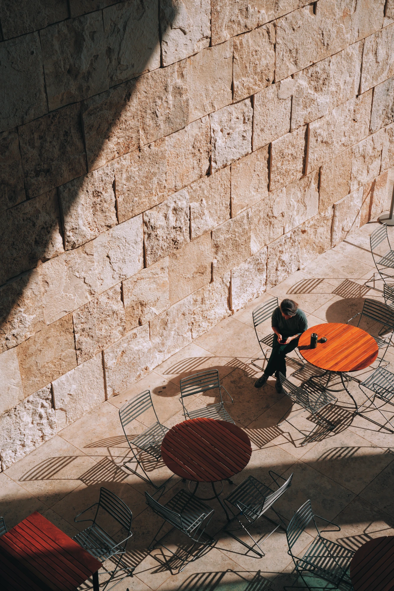 Hard sunlight casts table and chair shadows on a stone plaza beside a seated visitor.