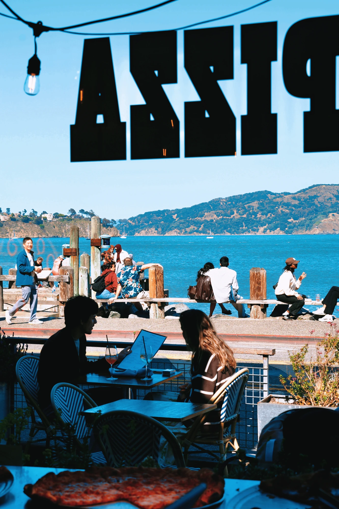 Diners sit beneath a pizza sign overlooking a waterfront.
